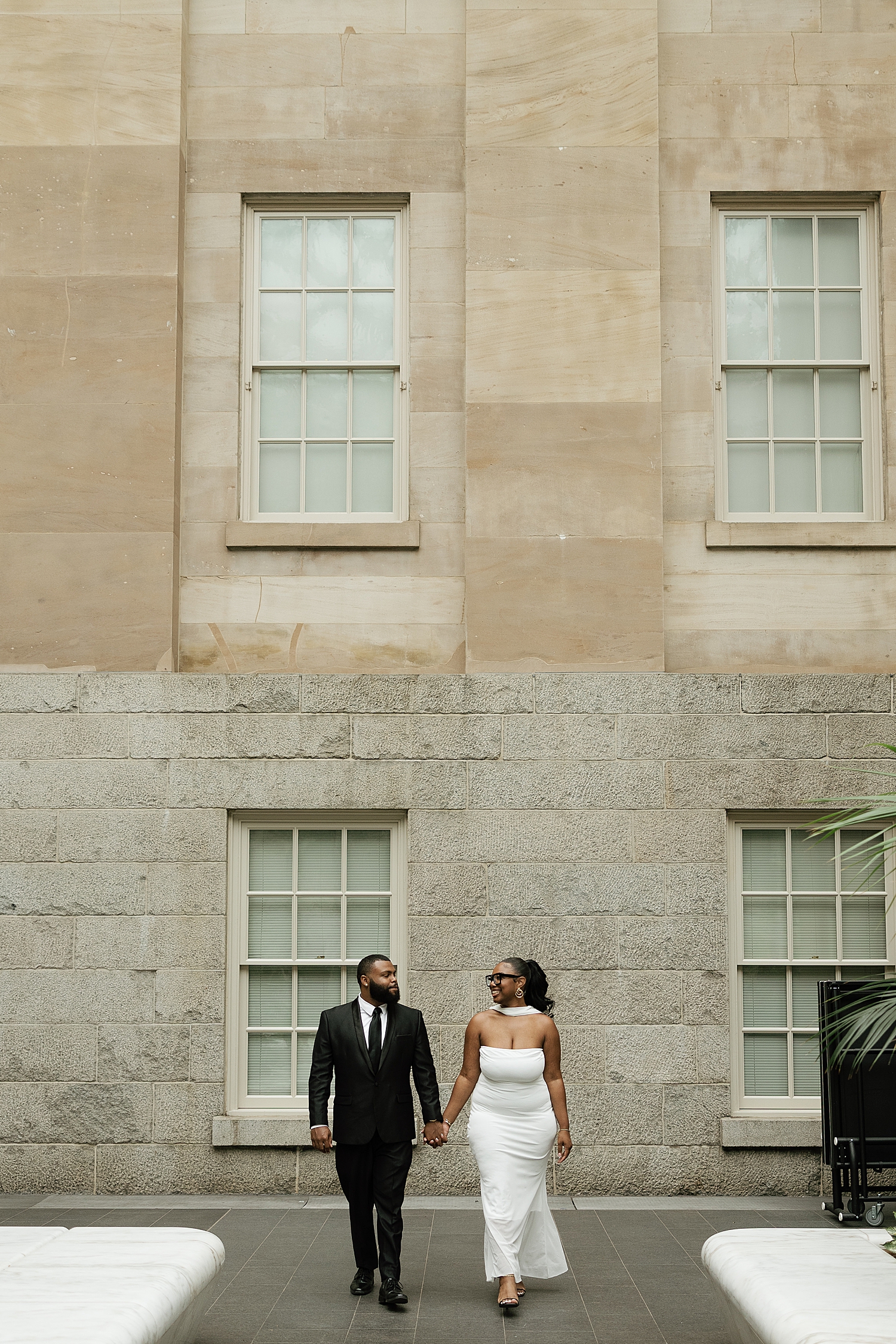 man and woman walk hand in hand in front of big stone building by Sharp Impressions Photo