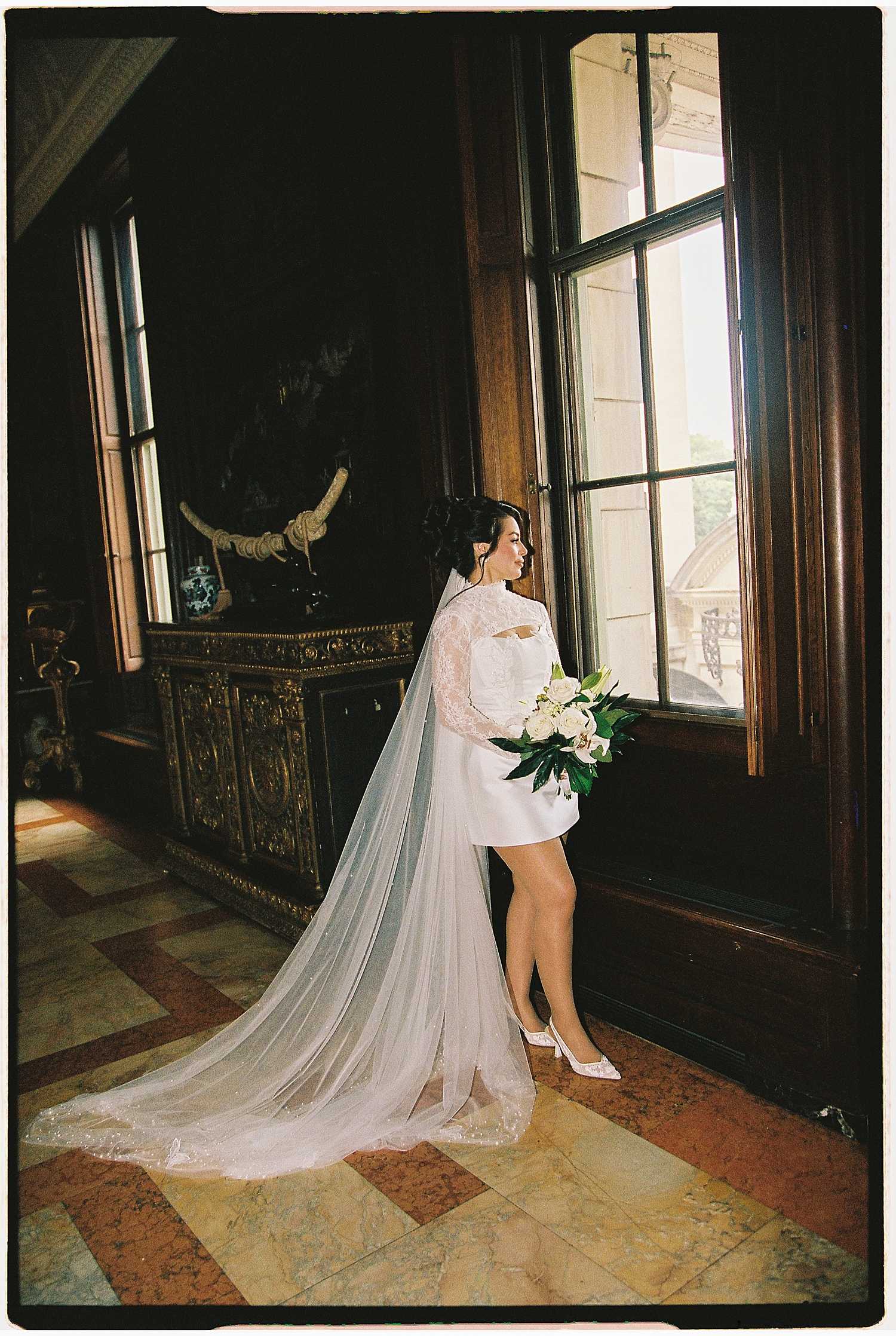 woman in white mini dress and veil stands by big window by Maryland wedding photographer